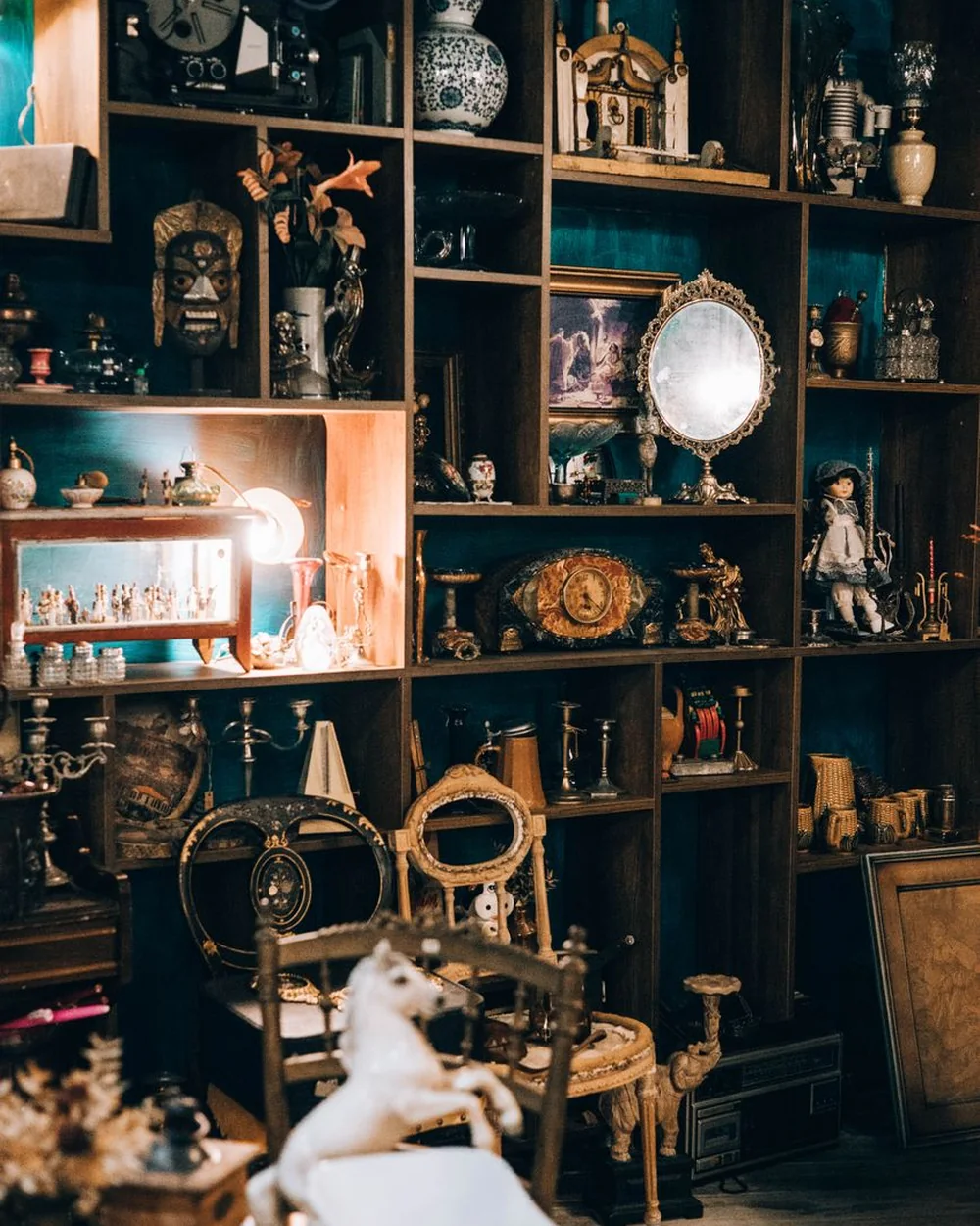 A dense wall of shelved antiques — clocks, dolls, mirrors, candelabra and curios (representative photograph of an antiques collection, not Primuseum's own shelves)